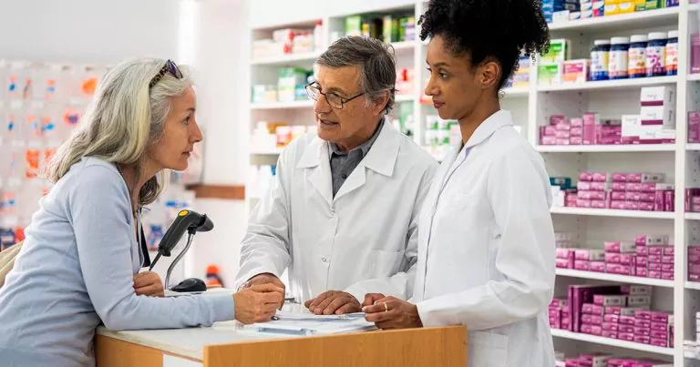 Woman consulting with two pharmacists at a pharmacy counter with shelves filled with drugs in the background.