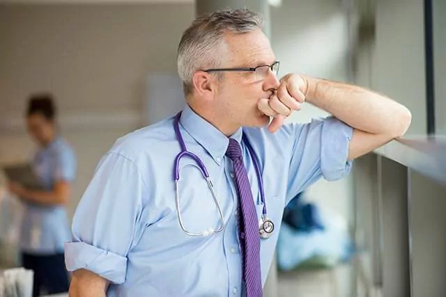 A male doctor in a blue shirt, and tie, with a stethoscope leans against a window, looking thoughtfully outside.