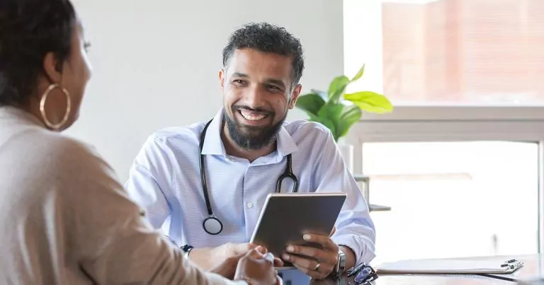 Doctor holding a tablet, listening to a patient in a well-lit consultation room.