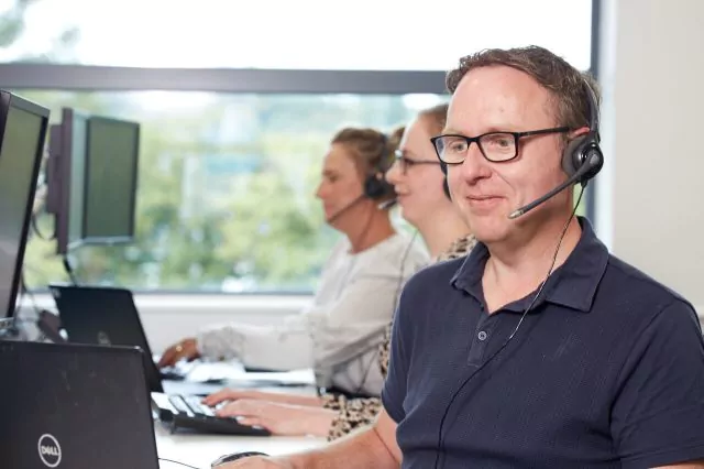 Smiling man with glasses wearing a headset and black zip-up jacket, looking at a laptop screen.
