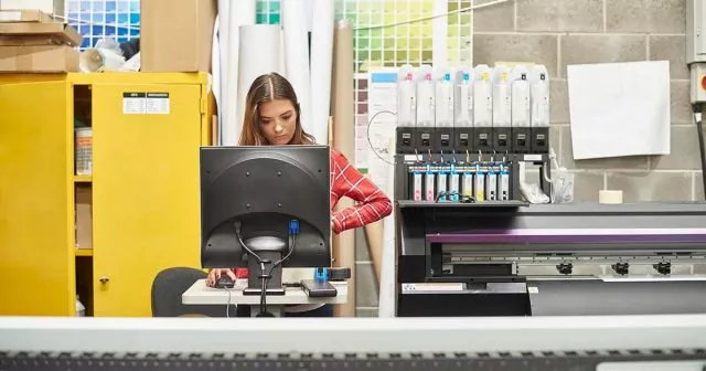 Woman in a plaid shirt standing by a printer in a print shop with printing equipment around her.