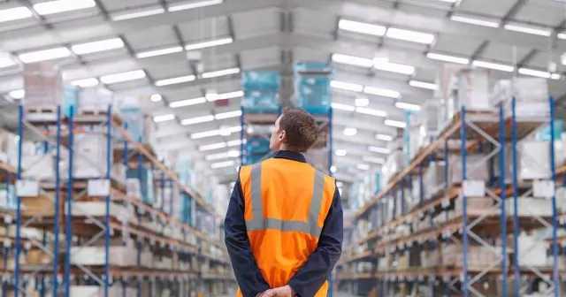 A person wearing an orange safety vest stands in a large warehouse, with shelves stacked high with boxes and pallets.