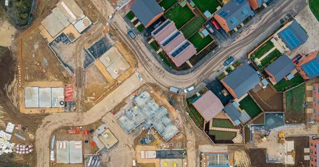 Aerial view of a residential construction site. Several houses are built with roofs installed, while others are just foundations. A curved road separates the completed houses from the construction area.