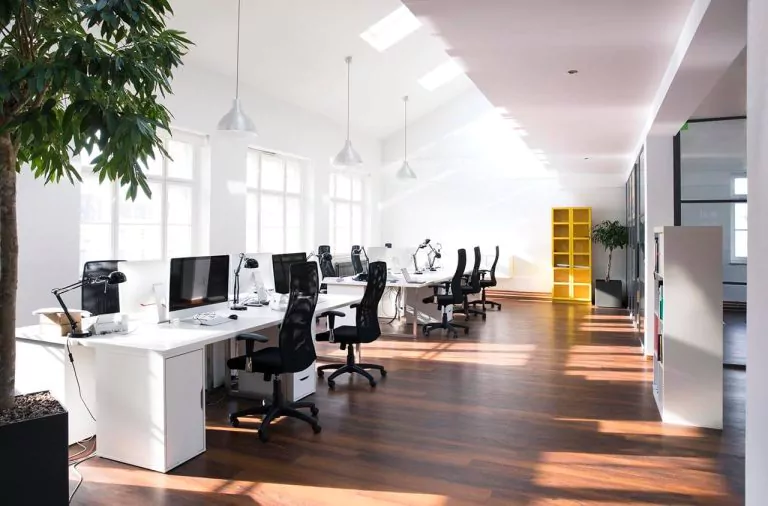 Empty office space with bare desks and chairs, large windows letting in natural light, and plain white walls.