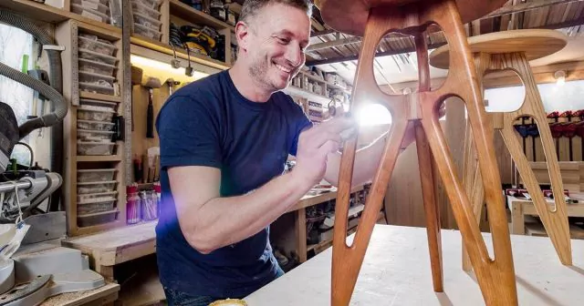 A man in a navy shirt is smiling as he polishes a wooden stool. With the workshop filled with tools and materials.