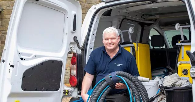 Man sitting at the back of an open white van with cleaning supplies, holding a large hose.