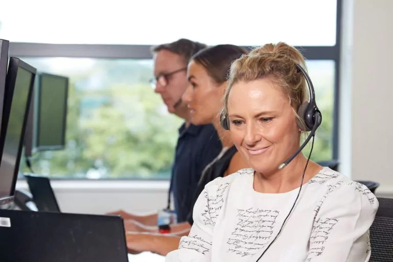 Smiling customer service advisor wearing a headset, seated at a desk in a bright office space.