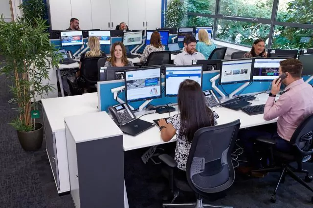 Group of office employees sat at their desks, in a brightly lit workspace. Some individuals are engaging with computers or phones.
