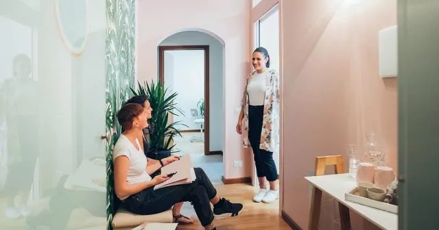 Three people in a hallway, two sitting, one standing and smiling at them near a plant.