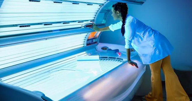 Person in a medical apron adjusting the lid of a tanning bed in a dimly lit room.