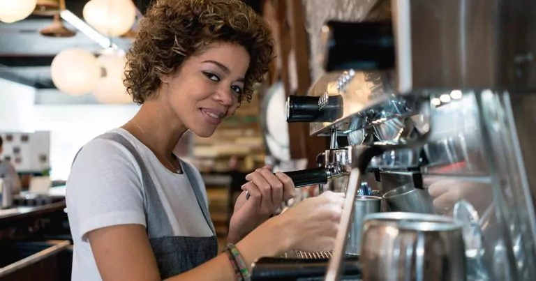 A person with curly hair is operating an espresso machine in a coffee shop. They are wearing a gray apron and smiling at the camera. There are shelves with items and round hanging lights in the background.