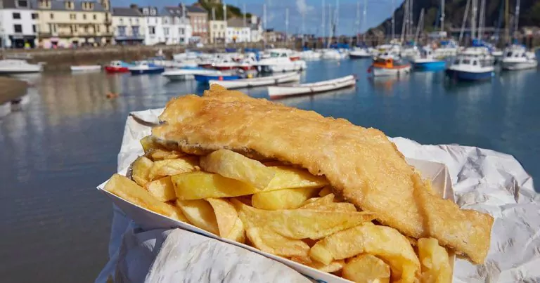 A serving of fish and chips sits on white paper in the foreground, with a harbor containing numerous boats and a row of buildings along the waterfront in the background under a clear blue sky.