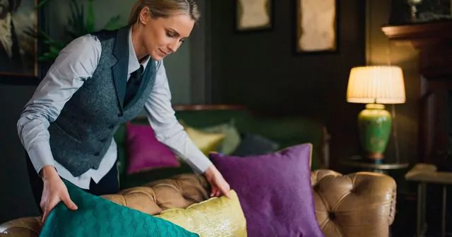 A person arranges colorful cushions on a brown leather sofa in a dimly lit, stylish room reminiscent of a chic hotel. A green lamp and framed artwork add flair.