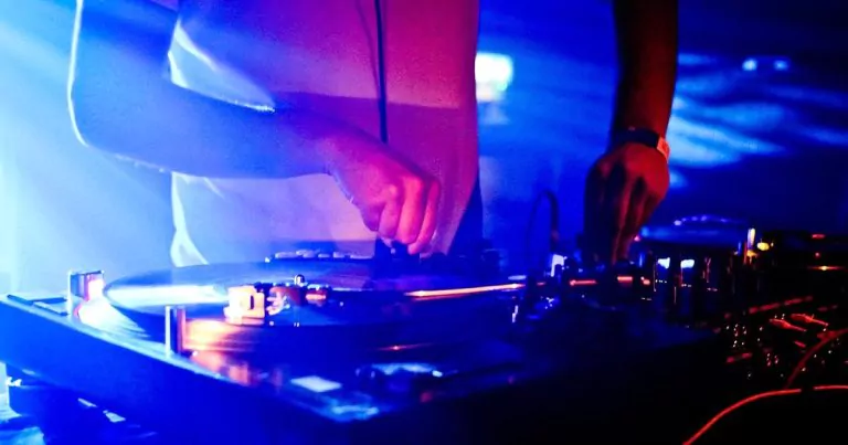 A DJ is mixing music on a turntable in a dimly lit environment with blue and pink lights. The image focuses on their hands adjusting records and controls, with various knobs and equipment visible.