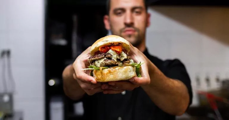 Man holding a large sandwich in a kitchen with grilled meat, cheese, and vegetables between a bun, background blurred.
