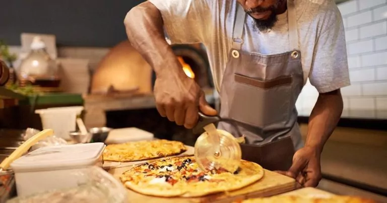 A person wearing an apron is cutting a freshly baked pizza with a pizza cutter. The pizza is topped with various ingredients. A brick oven is visible in the background along with more pizzas on the counter.