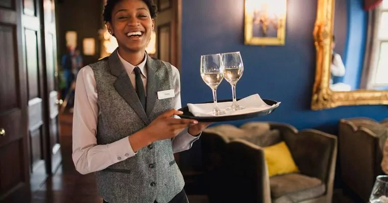 A person wearing a gray vest and white shirt holds a tray with two glasses of white wine. They are standing in a warmly lit room with blue walls and vintage decor, including a large gold-framed picture. The person is smiling.