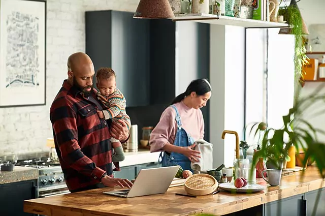 Man holding a child while using a laptop on a kitchen island, with a woman wiping her hands with a towel next to them.