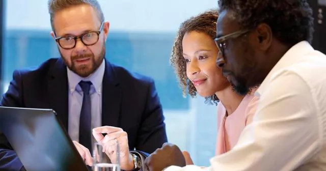 A group of three professionals speaking, all seated around a table looking at a laptop.