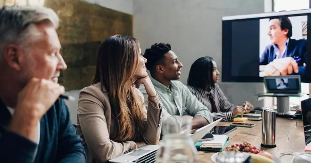 Group of four people in a meeting room with a video call on a large screen at the table.