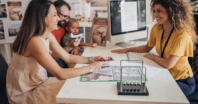 Travel agent showing information to a smiling family seated at a desk, with travel posters in the background.