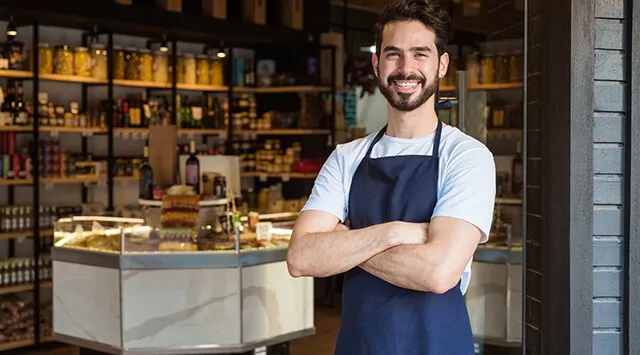 A man wearing a blue apron stands with his arms crossed in front of a deli or grocery store. Shelves filled with various products are visible in the background. He is smiling and standing in the entrance of the store.