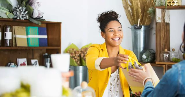Smiling female shop owner standing behind the counter in a retail store, handing a shopping bag to a customer.