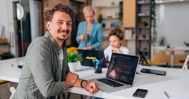 Man at a table with a laptop displaying code, in a casual workspace with two colleagues in the background.