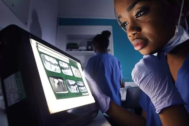 A dental professional in blue scrubs and gloves examines X-rays on a lightbox. Another person, also in scrubs, is in the background.