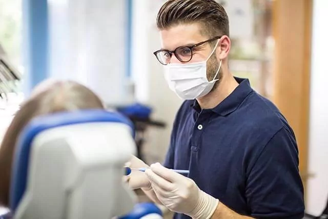 A person wearing glasses, a face mask, and gloves is sitting in a dental office, holding dental instruments. Another person, partially visible, is seated in a dental chair facing away from the camera.