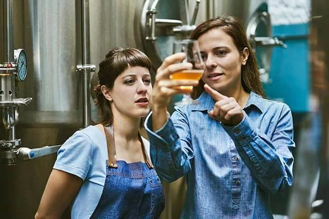 Two women stand in a brewery, discussing brewery insurance. One holds a glass of beer, observing its color while pointing at it with interest. Both wear blue clothing as large metal brewing tanks loom in the background.