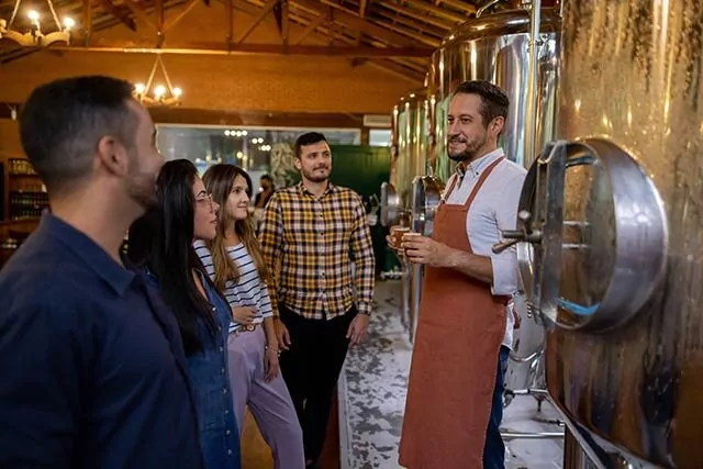 A man wearing an apron is giving a tour to a group of five people in a brewery. He stands in front of large stainless steel tanks, explaining the brewing process amid wooden beams and soft lighting.