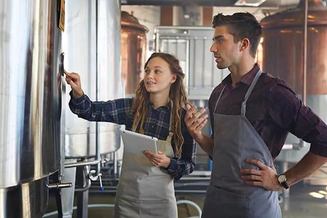 Two people in aprons stand in a brewery. One holds a tablet and points at a metal container, while the other stands with a hand on their hip. Industrial equipment is visible in the background.
