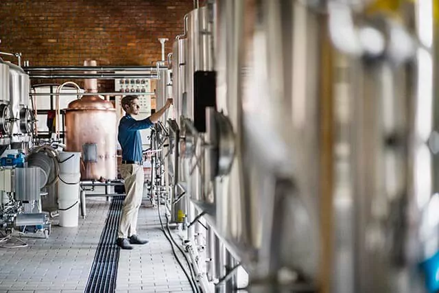 Person operating or inspecting brewing equipment in a spacious brewery with tanks and a copper vat.