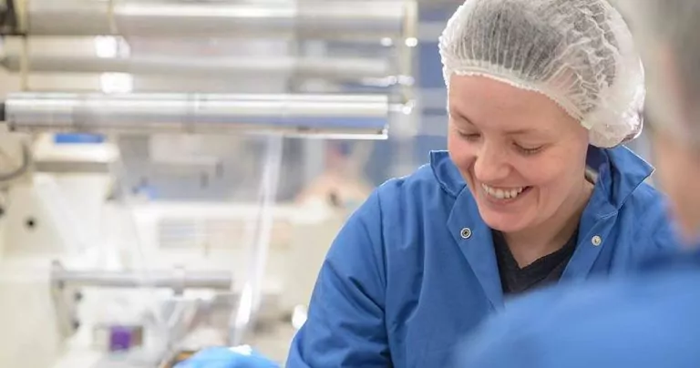 Worker smiling in a food manufacturing facility, wearing a lab coat and hairnet with equipment in the background.
