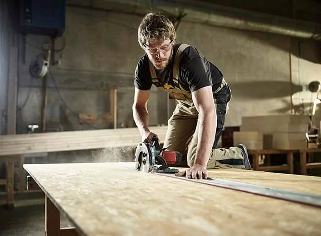Person cutting a wooden board with a circular saw in a workshop, dust visible in the air.