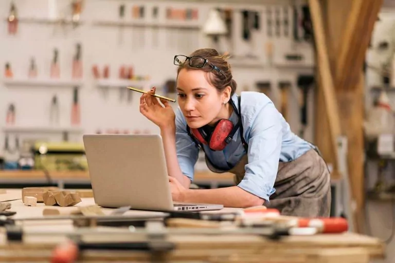 Woman working on a laptop in a workshop, holding a pencil with equipment and tools around her.
