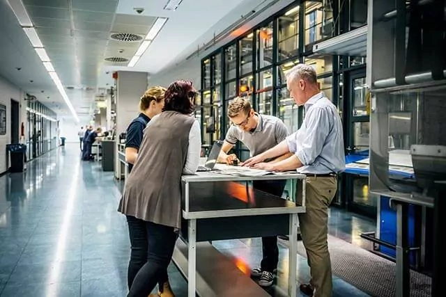 Four people stand around a table in a brightly lit hallway, engaged in discussion. They are examining engineering blueprints. Shelves filled with books or files line the wall in the background.