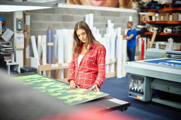 A woman works at a table with green and black patterned material. There is printing equipment in the background.