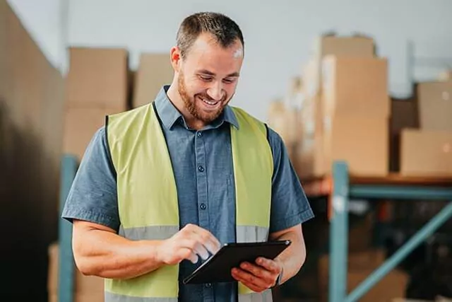 Man in a warehouse using a tablet, with stacks of boxes and metal shelving in the background.