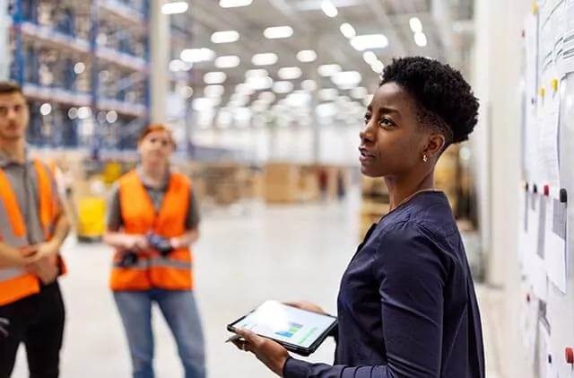 Person with tablet reviewing manufacturing processes in a warehouse, with two workers in the background.