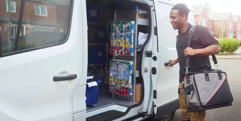Trade worker beside an open van door, with tools and stock visible in the van, smiling with houses in the background.