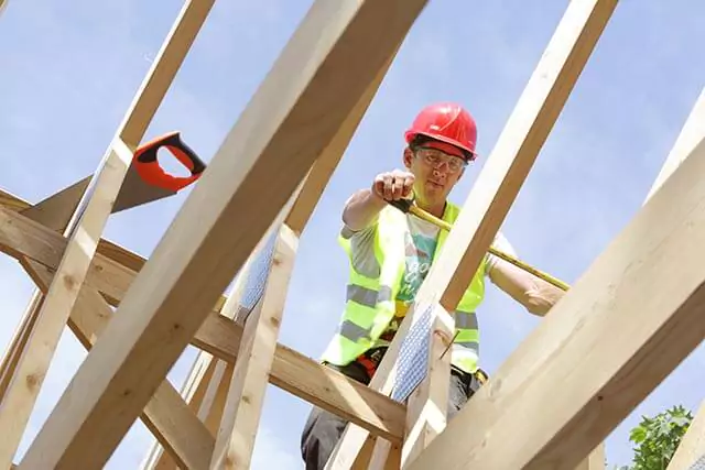 Worker in a red hard hat measuring a wooden beam on a roof, with a clear blue sky backdrop.