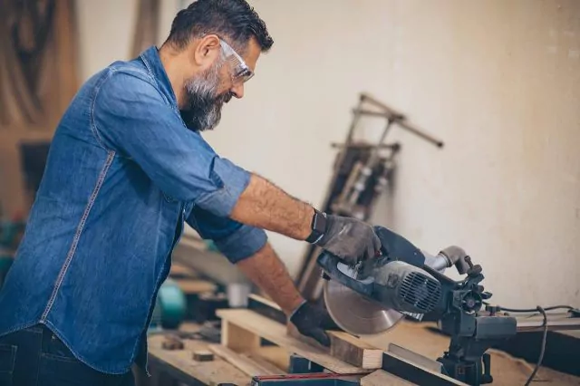 Wood worker using a miter saw to cut wood in a workshop, surrounded by tools.
