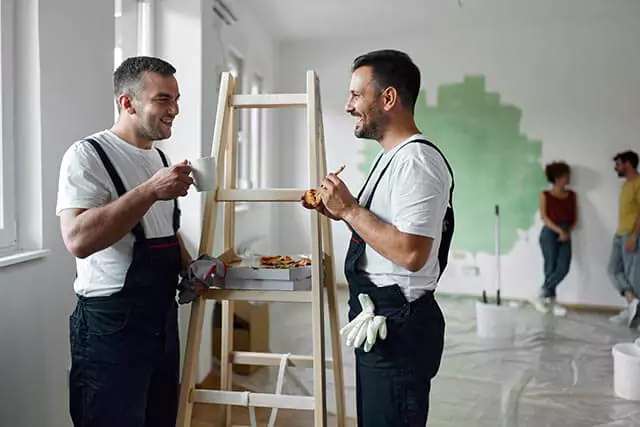 Two men in work overalls stand beside a wooden ladder, holding slices of pizza and a mug, and smiling at each other. In the background, two people near a partially painted wall appear to be talking. Paint supplies are scattered around the room.
