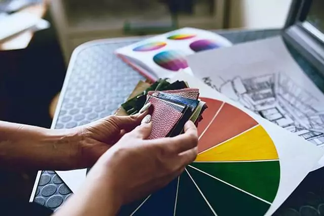 A person holds a selection of fabric swatches in their hands, sitting on a dark table with a color wheel and an architectural sketch nearby. The scene suggests a design or planning process.