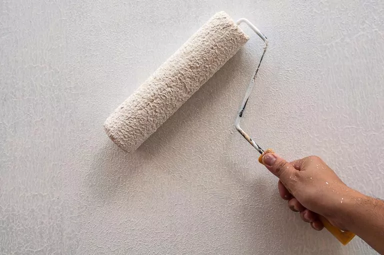 Person painting a wall white using a paint roller. The wall is textured and paint splatters are visible.