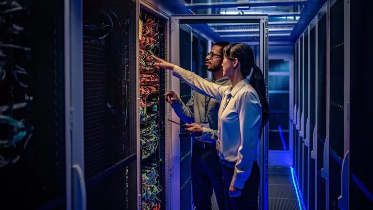 Two people are standing in a dimly lit server room. They are examining and pointing at the cables in a network server rack. The room has a modern appearance, with glowing blue lights on the floor.