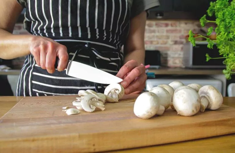 Chef slicing mushrooms in a kitchen, with herbs and kitchen appliances in the background.