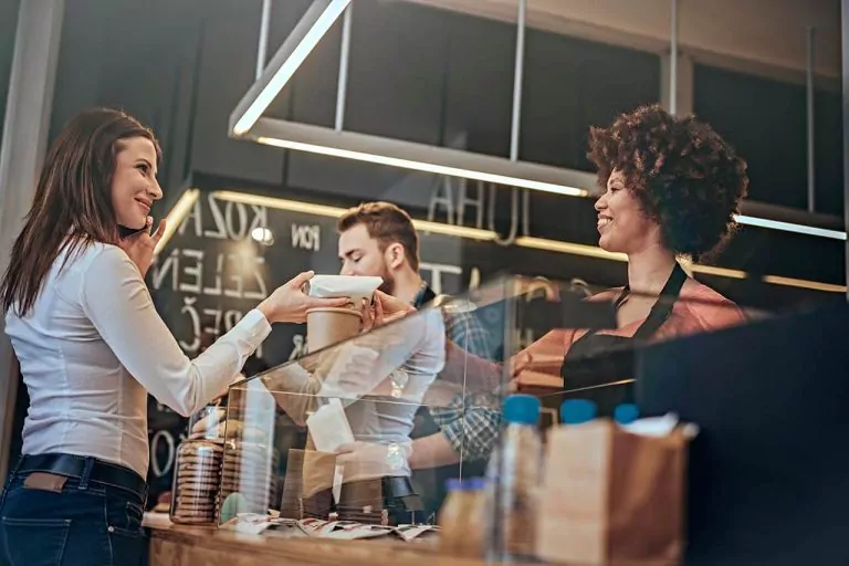 Woman at café counter receiving a wrapped dessert or pastry from a smiling barista, a man preparing a drink in the background.
